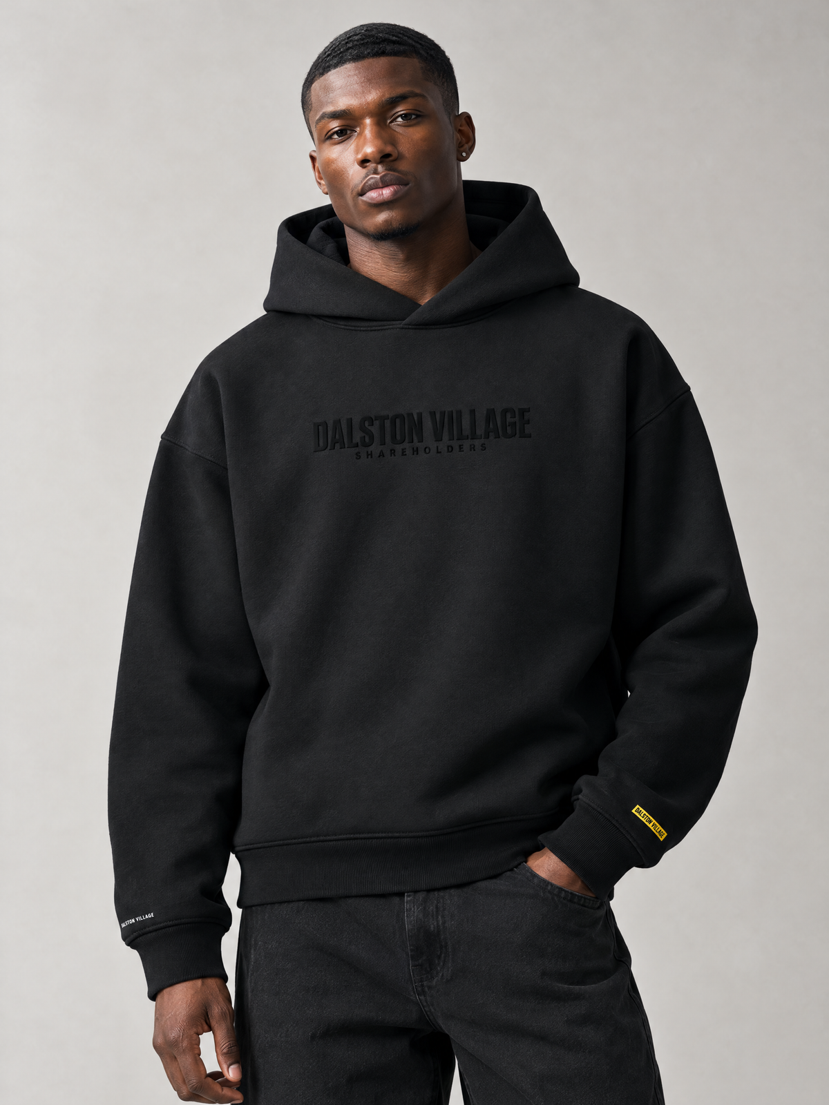 AI-generated editorial styling mockup of a model wearing the Dalston Village Shareholders Black Phase heavyweight hoodie; black-on-black chest wordmark and yellow cuff mark. Illustrative styling, not physical fit proof.