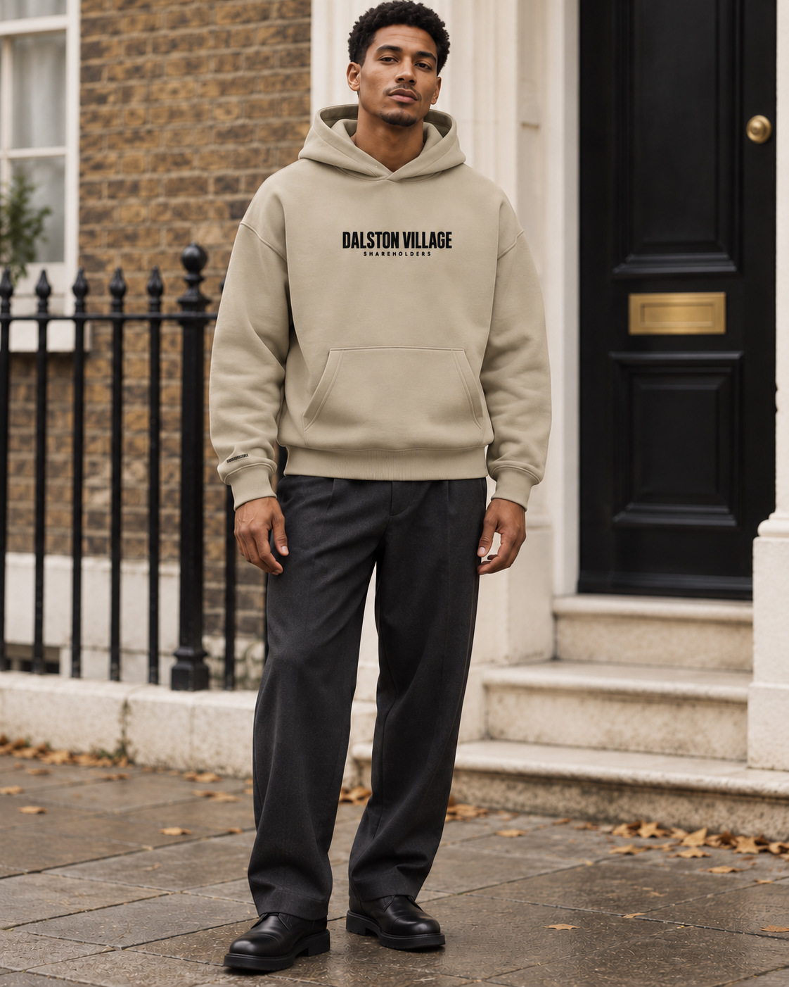 AI-generated editorial mockup of a model wearing the sand Dalston Village Shareholders hoodie with charcoal trousers.